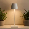 A white table lamp with a gray shade sits on a white surface between two potted plants. Náhled obrázku produktu