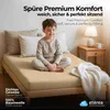 A young boy sitting on a bed with taupe bedding, with a bedside table and lamp in the background. Produktbild-Vorschau