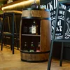 A wooden barrel-shaped bar cabinet with bottles of alcohol displayed inside, set in a bar or home setting with stools and a textured wall in the background. Náhled obrázku produktu