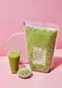 A bag of green powder, a glass of green liquid with a straw, and a small bowl of green seeds are arranged on a pink background. Produktbild-Vorschau