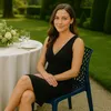 A woman sits at a table in a garden setting, wearing a black dress and sitting on a dark blue chair. Produktbild-Vorschau