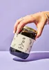 A hand holding a jar of syrup against a purple background. Produktbild-Vorschau