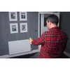 A man in a red plaid shirt is using a yellow and black infrared thermometer to measure the temperature of a white radiator in a room with patterned wallpaper. Produktbild-Vorschau