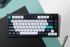 A black mechanical keyboard with white and dark blue keycaps is placed on a background with geometric shapes in black, teal, and white. Voir les images du produit