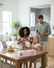A family is gathered around a table in a bright, modern home. The mother is styling her daughter's hair with a hair tool, while the father stands behind them, looking at his phone. There's a laptop, fruit, and a product box on the table. Produktbild-Vorschau