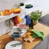 A kitchen counter scene with various colored cloths, oranges, and a plant. Produktbild-Vorschau