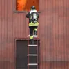 A firefighter in full gear is climbing a ladder up the side of a wooden building towards a window with flames visible inside. Voir les images du produit