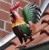 A colorful metal rooster figurine is perched on a tiled roof, next to a gutter. Produktbild-Vorschau