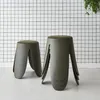 Two khaki-colored stools with green upholstered seats are placed on a light gray floor in front of a white grid-patterned wall. A green plant hangs from the top right corner. Produktbild-Vorschau