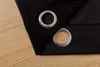 A close-up view of two grommets on a black curtain, with a wooden floor visible below. Produktbild-Vorschau