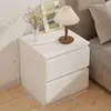 A white bedside table with two drawers is placed next to a sofa. A lamp and a vase with flowers are on top of the table. Produktbild-Vorschau