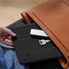 A hand is holding a black tablet case with a silver USB hub and a key tucked inside, placed on a person's lap, with a brown leather bag partially covering the tablet. Anteprima dell’immagine prodotto
