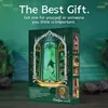 A product box for a DIY Book Nook is displayed on a wooden surface with a blurred green background. Náhled obrázku produktu