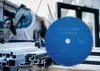 A close-up of a blue circular saw blade with text, placed in front of a pile of aluminum profiles and a miter saw in the background. Voir les images du produit
