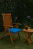 A wooden chair with a blue cushion and a small wooden table with two drinks on it, set on a grassy lawn with a green hedge in the background. Produktbild-Vorschau