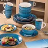 A table setting with blue ceramic dishes, including plates, bowls, and mugs, filled with food and drinks. Náhled obrázku produktu