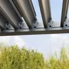 Close-up view of the metal structure of a pergola roof, with blurred green trees and blue sky in the background. Voir les images du produit