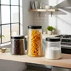 Three glass storage jars with black lids are displayed on a wooden shelf in a kitchen setting. Produktbild-Vorschau