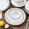 A set of white ceramic plates and bowls with a black rim, arranged on a brown surface. Lemons and a glass of water are also visible. Produktbild-Vorschau