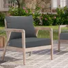An outdoor chair with dark gray cushions sits on a patio with a stone wall and flowering bushes in the background. Náhled obrázku produktu
