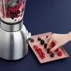 A hand is adding berries to a tray next to a blender filled with berries. Voir les images du produit