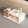 A modern coffee table with a white high-gloss finish and a wood-tone section, placed on a gray carpet in a living room setting. It features a glass shelf and is decorated with a bowl of fruit and a vase of flowers. Produktbild-Vorschau