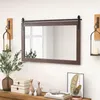 A dark brown rectangular mirror with a sliding barn door hardware is mounted on a white wall above a wooden console table. The table is decorated with various items, including a vase with greenery, decorative boxes, cat figurines, and books. Podgląd zdjęcia produktu