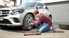 A man is using a cordless impact wrench to tighten the lug nuts on a car wheel. A spare tire is visible next to the car. Náhľad obrázku produktu
