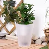 A frosted white planter with a green plant sits on a wooden deck, surrounded by rocks and ivy on a trellis. Produktbild-Vorschau