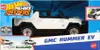 The image shows the packaging of a Hot Wheels Pull-Back Speeders GMC Hummer EV toy car. The packaging features a dynamic blue background with graphics suggesting speed and motion. The toy car itself is prominently displayed in white with black accents and  Anteprima dell’immagine prodotto