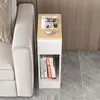 A narrow side table with a drawer and open shelf, placed next to a sofa. The table has a magazine and a cup of coffee on top, and books on the shelf. Náhled obrázku produktu