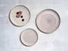 Three ceramic plates are arranged on a light gray textured surface. The largest plate is on the right, a medium-sized plate with figs and blackberries is on the top left, and a small plate is on the bottom left. Produktbild-Vorschau