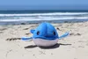 A plush whale toy is sitting on a sandy beach with the ocean in the background. Produktbild-Vorschau