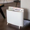 A white magazine rack with magazines inside, placed on a wooden floor next to a sofa. Podgląd zdjęcia produktu