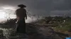 A lone figure in a straw hat stands on a dirt path, looking out over a misty, war-torn landscape with scattered flags and debris. Produktbild-Vorschau