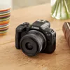 A black Canon EOS R100 camera with a lens attached is placed on a wooden table, next to a colorful striped mug and a blurred vase with greenery. Voir les images du produit