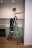 A woman is cleaning a kitchen cabinet while standing on a stepladder. Anteprima dell’immagine prodotto