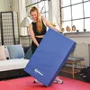 A woman is holding a blue foldable gym mat in a living room setting. Produktbild-Vorschau