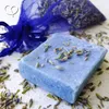 A bar of blue lavender soap with dried lavender sprigs on top, surrounded by loose dried lavender and a blue mesh bag filled with lavender. Produktbild-Vorschau