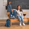 Two young girls are in a classroom setting with a blackboard behind them. One girl is writing on the blackboard, and the other is sitting on a bench next to a large backpack. Another backpack is on the floor. Voir les images du produit