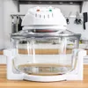 A white halogen oven with a glass bowl sits on a wooden countertop in a kitchen setting. Various kitchen utensils are visible on a shelf in the background. Podgląd zdjęcia produktu