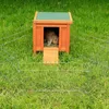 A rabbit is inside a wooden hutch with a ramp leading out, surrounded by a wire fence in a grassy yard. Produktbild-Vorschau