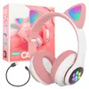 A pink wireless headset with cat ears and LED lights, shown with its charging cable and packaging in the background. Náhľad obrázku produktu