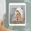 A woman with a towel on her head is looking into a lighted mirror, touching her face. The mirror displays a digital clock and has a feature indicator. Produktbild-Vorschau