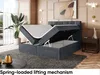A dark gray upholstered bed frame with its storage compartment open, revealing a spring-loaded lifting mechanism. The bed is in a stylish bedroom setting with decorative elements. Produktbild-Vorschau