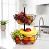 A two-tiered black metal fruit basket filled with various fruits, placed on a white countertop in a kitchen setting. Produktbild-Vorschau