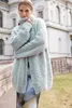 A woman wearing a light blue fuzzy cardigan stands in front of a building. Produktbild-Vorschau