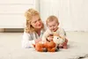 A woman and a baby are lying on a rug, playing with a stuffed deer toy. Produktbild-Vorschau