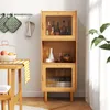 A wooden cabinet with glass doors, filled with bottles and kitchenware, stands next to a table with fruit. Produktbild-Vorschau