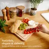 A wooden cutting board with bread, cheese, grapes, and tomatoes on a wooden countertop, with kitchen items in the background. Produktbild-Vorschau